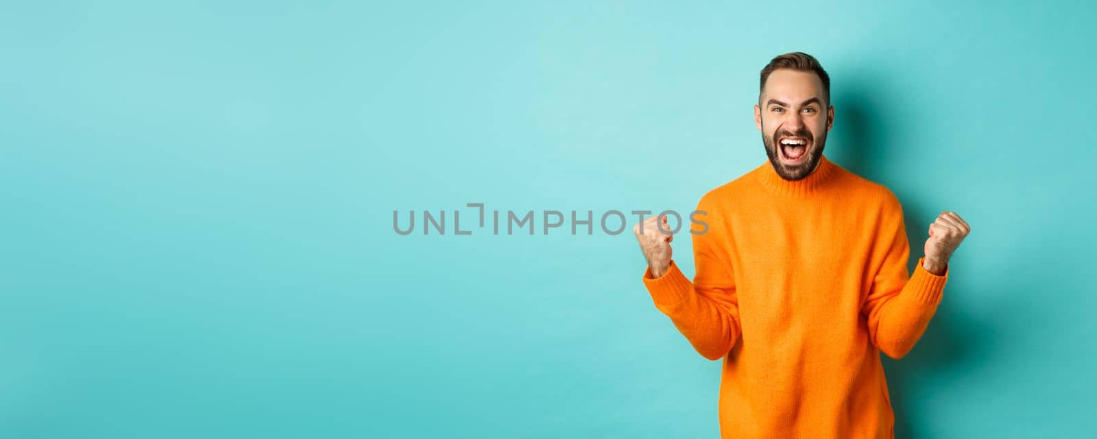 Excited man celebrating victory, rejoicing and making fist pump gesture, winning and looking satisfied, saying yes, achieve goal, standing over light blue background.