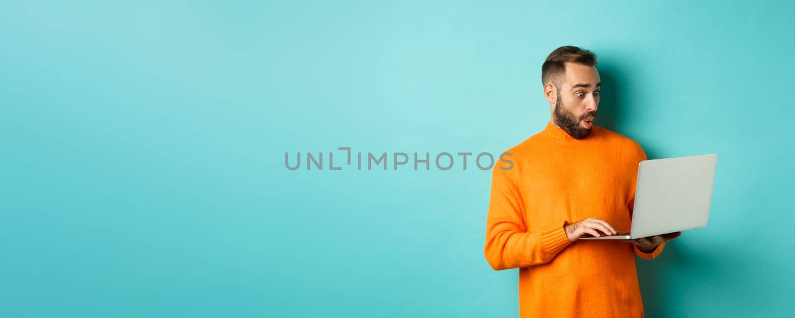 Freelance and technology concept. Image of handsome adult man staring at laptop with amazement, shopping online or working remote, standing over turquoise background by Benzoix