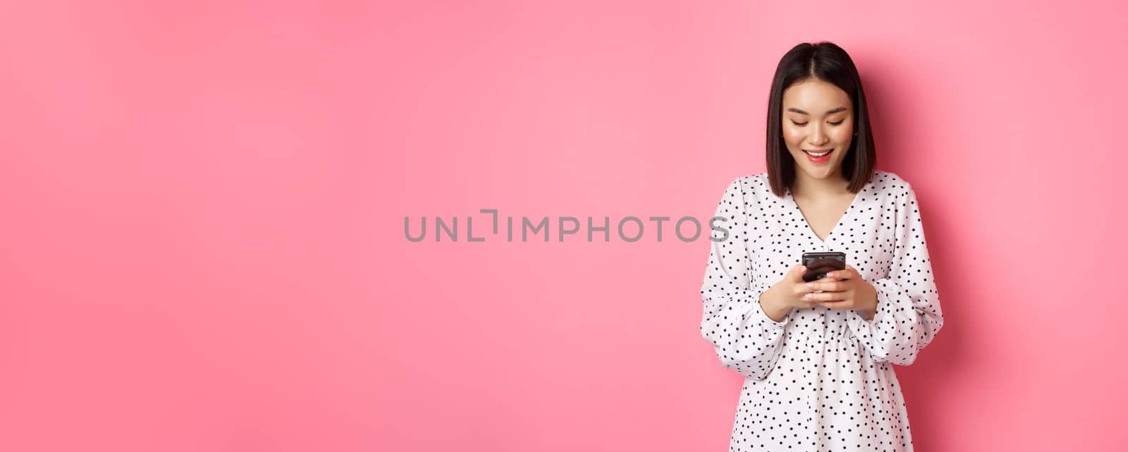 Beautiful asian lady reading message and smiling, using mobile phone, standing in cute dress against pink background.