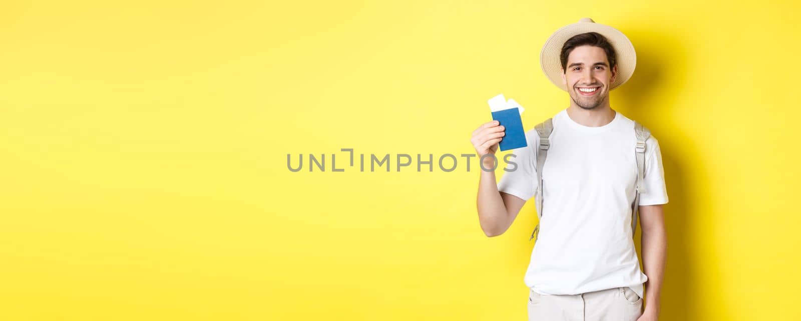 Tourism and vacation. Young smiling tourist showing passport with tickets, going on a trip, standing against yellow background by Benzoix