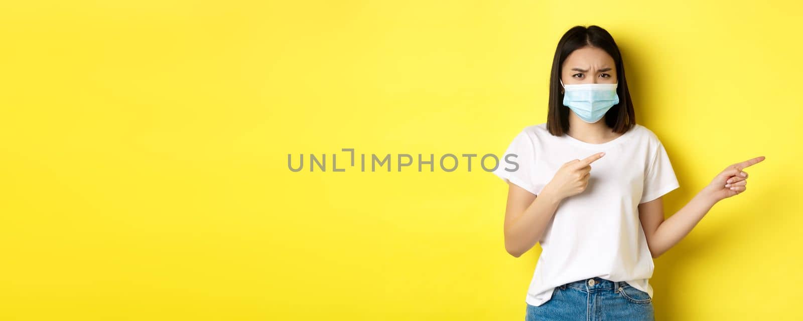 Covid-19, pandemic and social distancing concept. Disappointed asian girl in medical mask, frowning upset and pointing fingers right at logo, standing over yellow background by Benzoix