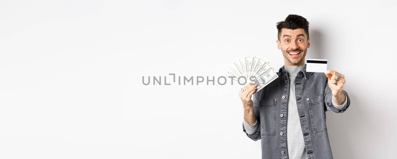Image of handsome guy holding dollar bills but suggesting use plastic credit card, smiling friendly at camera, standing on white background by Benzoix