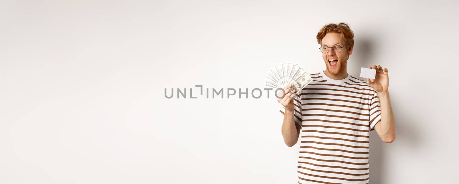 Shopping and finance concept. Cheerful handsome redhead man showing plastic credit card and cash, smiling amazed, white background by Benzoix