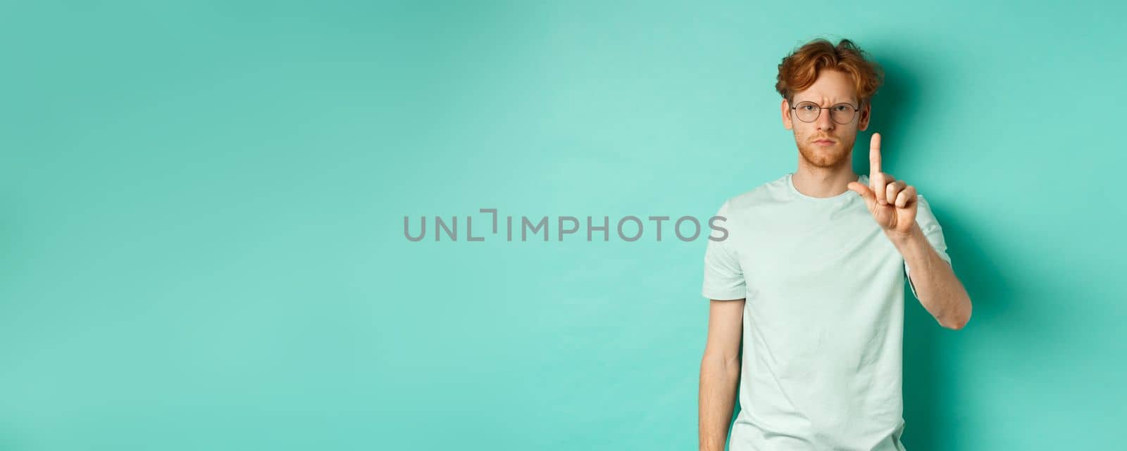 Angry and serious young man with red hair, wearing glasses, showing stop gesture, telling no, shaking finger with disapproval, standing over mint background by Benzoix