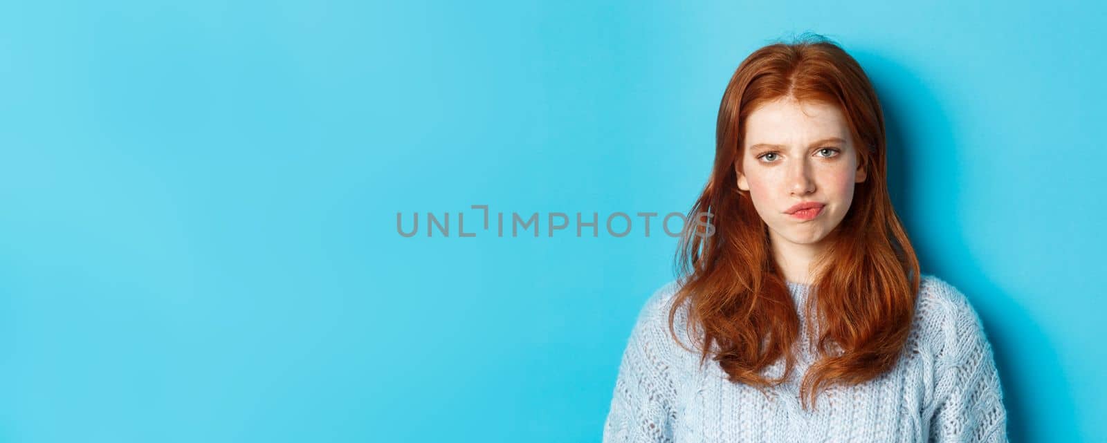 Disappointed teenage girl with red hair, frowning and smirking displeased, looking judgemental, standing against blue background.