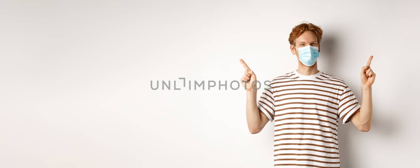 Covid, virus and social distancing concept. Handsome young man with red hair, wear face mask and pointing sideways at two promo offers, white background.