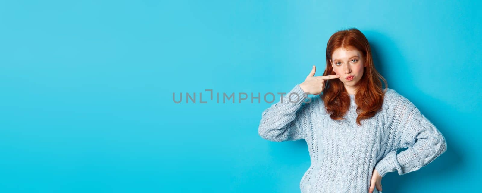 Cute redhead girl in sweater poking her cheek, staring at camera sassy, standing over blue background.