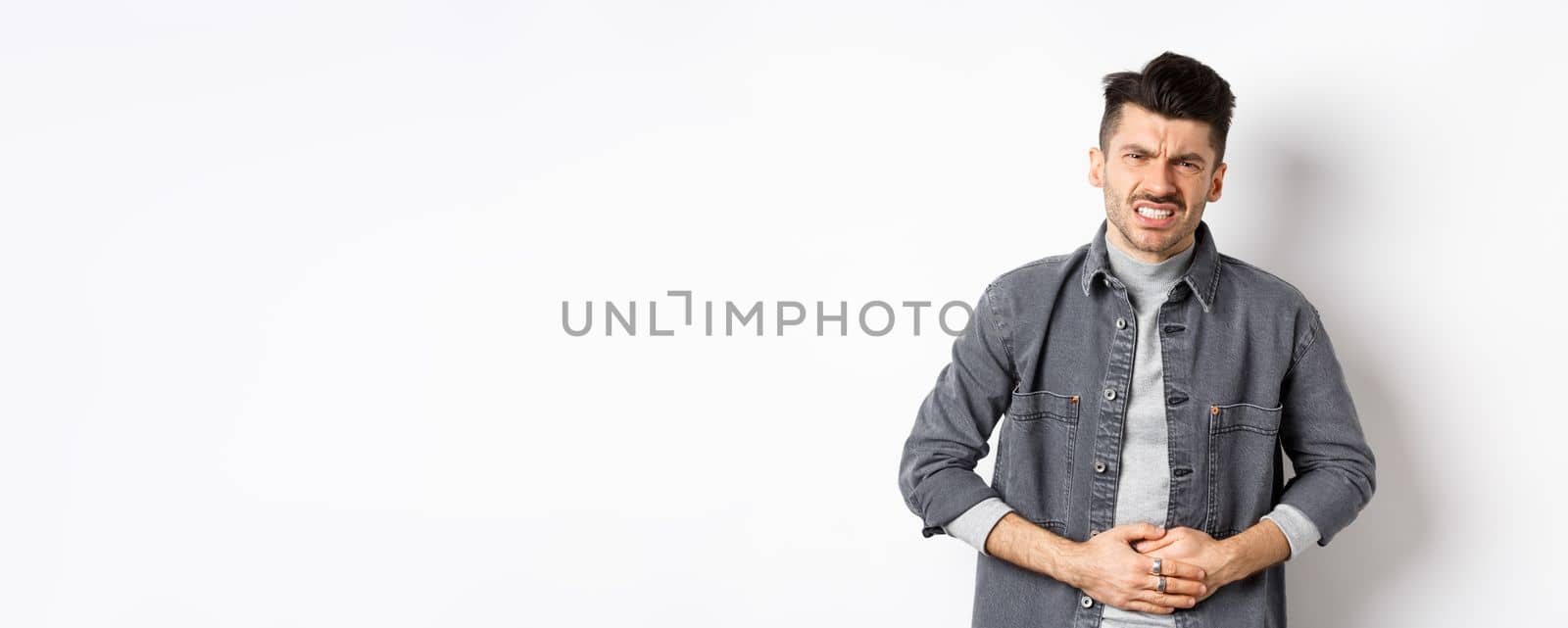 Man with stomach ache frowning and touching belly, having food poison, suffer diarrhea, standing on white background by Benzoix