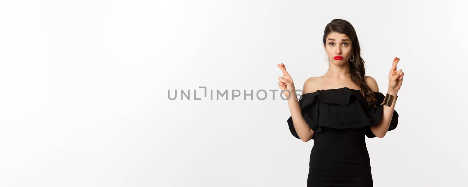 Fashion and beauty. Hopeful silly woman in black dress making wish, holding fingers crossed for good luck, standing over white background.