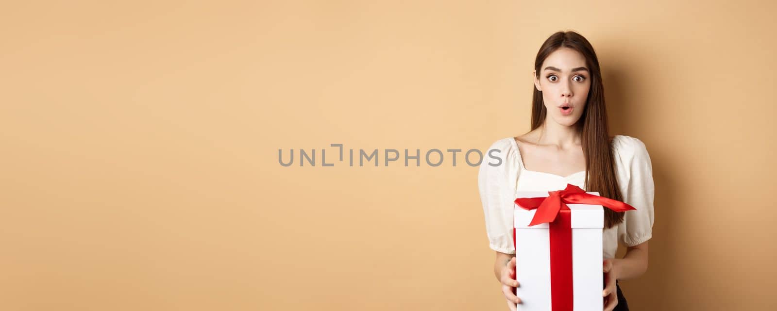 Image of surprised girl receive gift on Valentines day, looking with disbelief and holding present, standing on beige background.