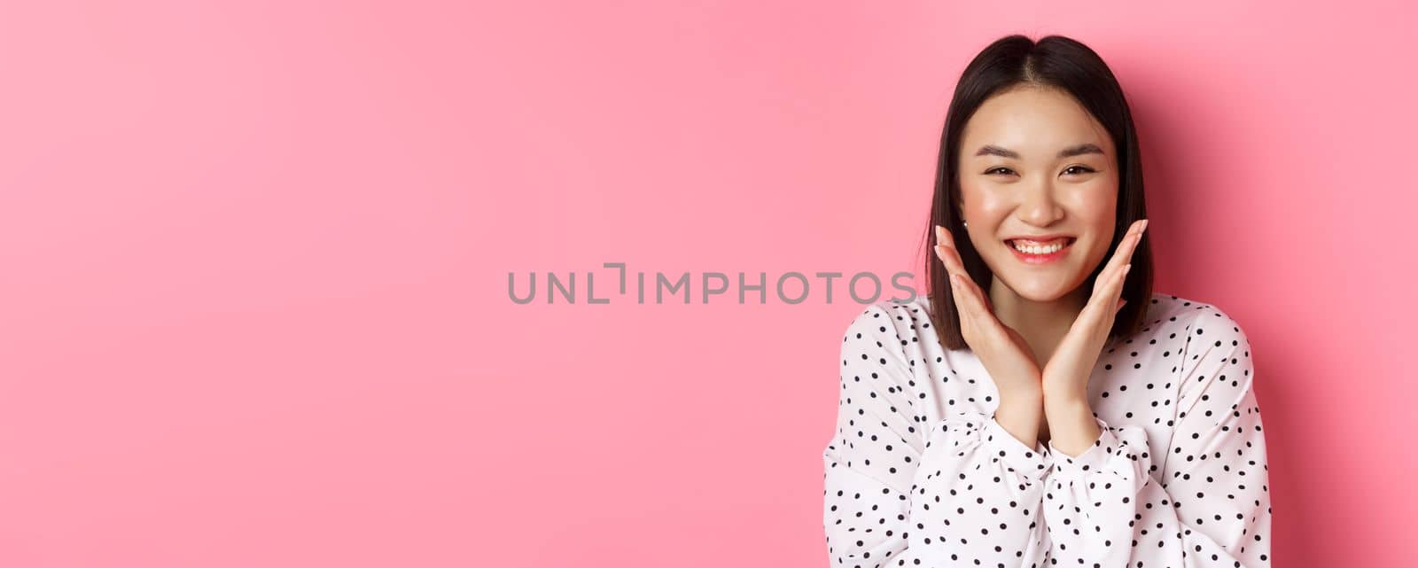 Beauty and skin care concept. Close-up of cute asian woman showing clean perfect face and smiling, looking happy at camera, standing over pink background by Benzoix
