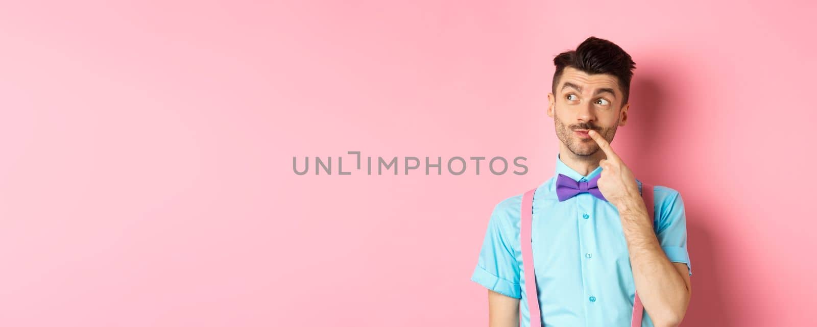 Image of handsome funny man in bow-tie looking thoughtful left and touching lip, thinking what to choose, making decision, standing over pink background by Benzoix