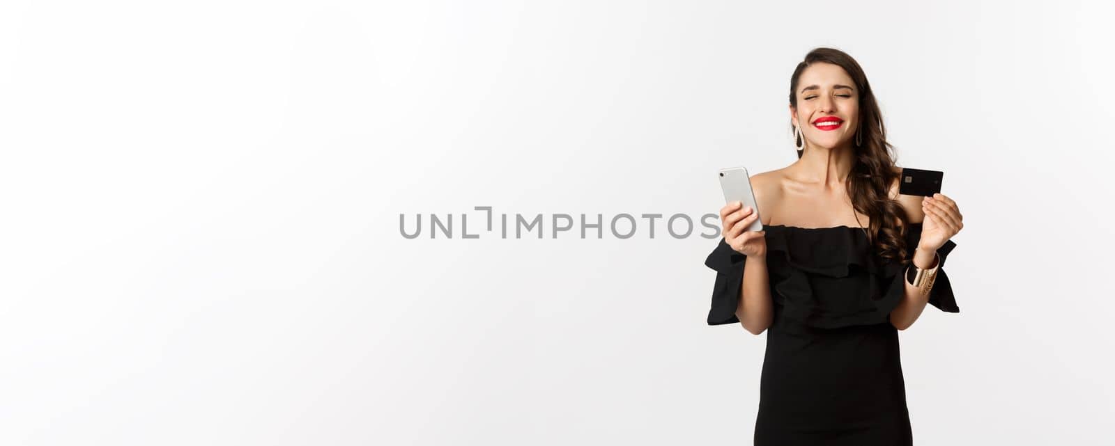 Online shopping concept. Fashionable woman in black dress, holding credit card with smartphone, looking satisfied, standing over white background.