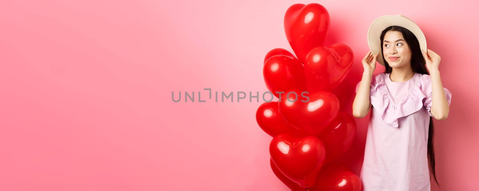 Tender and romantic asian teen girl wearing straw hat and dress on date, standing near valentines day heart balloons and looking aside with dreamy smile, pink background.
