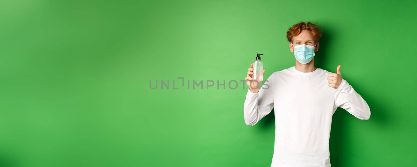 Covid-19, virus and social distancing concept. Smiling young man with red hair, wearing face mask for protection from corona, showing thumb-up and hand sanitizer, green background by Benzoix