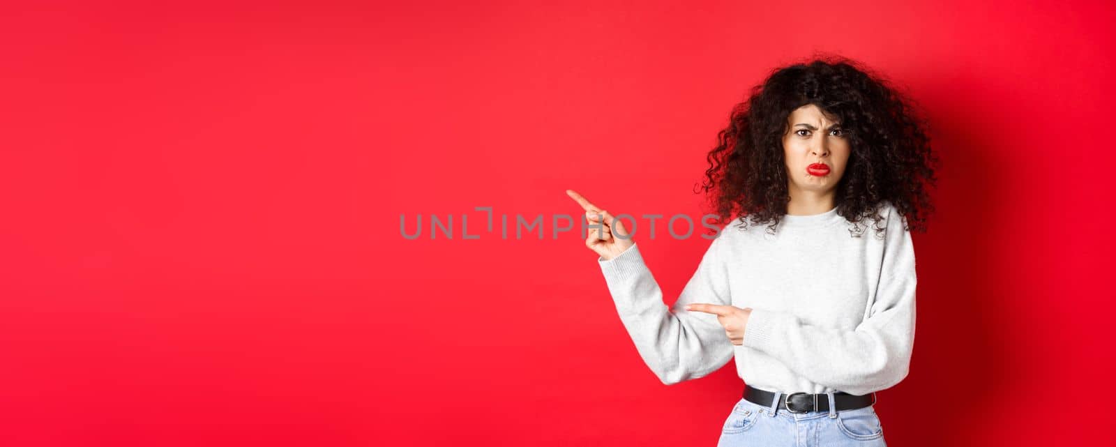 Disappointed funny woman, frowning and complaining, pointing fingers left at empty space and arguing, looking displeased, red background.