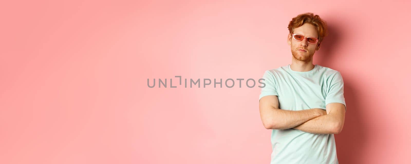 Tourism and vacation concept. Sassy redhead guy in sunglasses, cross arms on chest and looking serious at camera, standing over pink background.