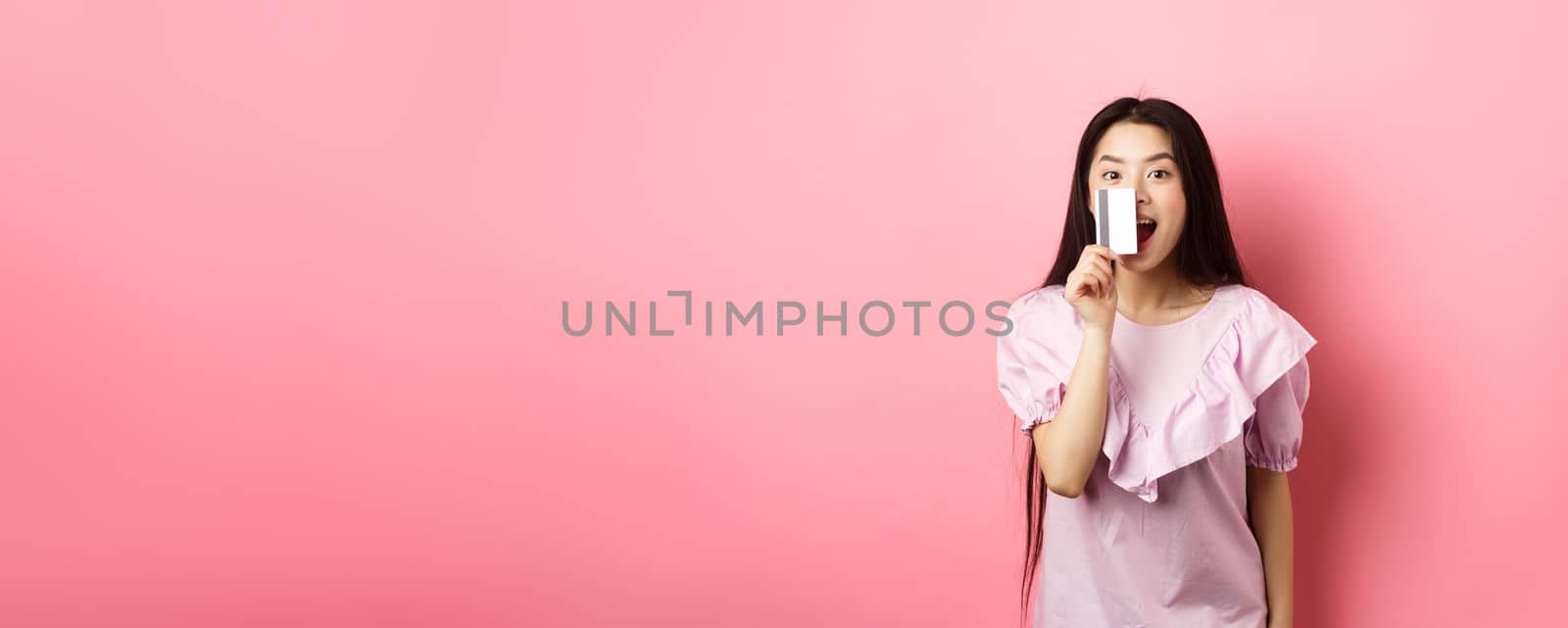 Excited asian girl showing plastic credit card and smiling, going on shopping, standing against pink background by Benzoix