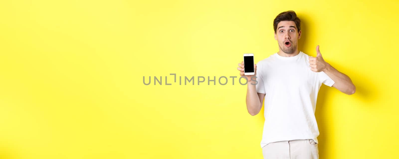 Impressed guy showing awesome smartphone app on screen and thumb-up, gasping amazed, standing against yellow background.