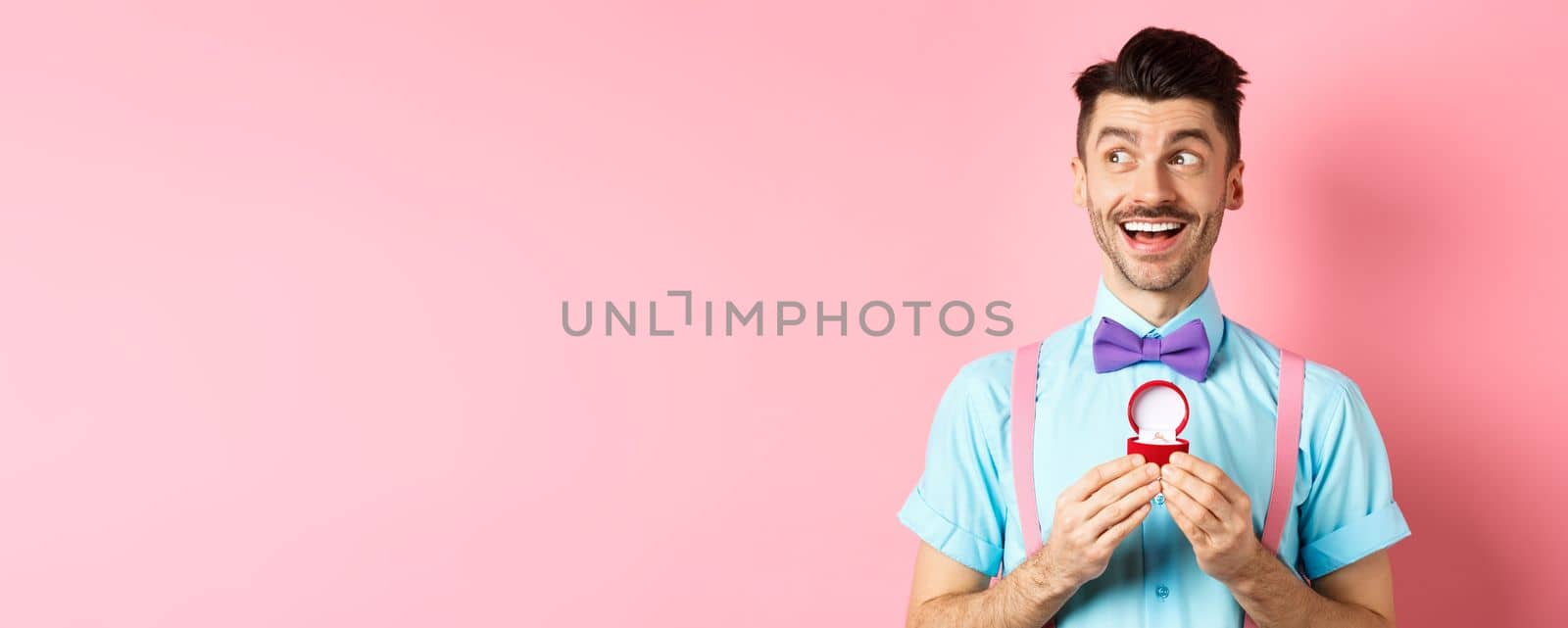 Valentines day. Romantic handsome man looking dreamy and smiling, showing engagement ring for his lover, standing happy over pink background.