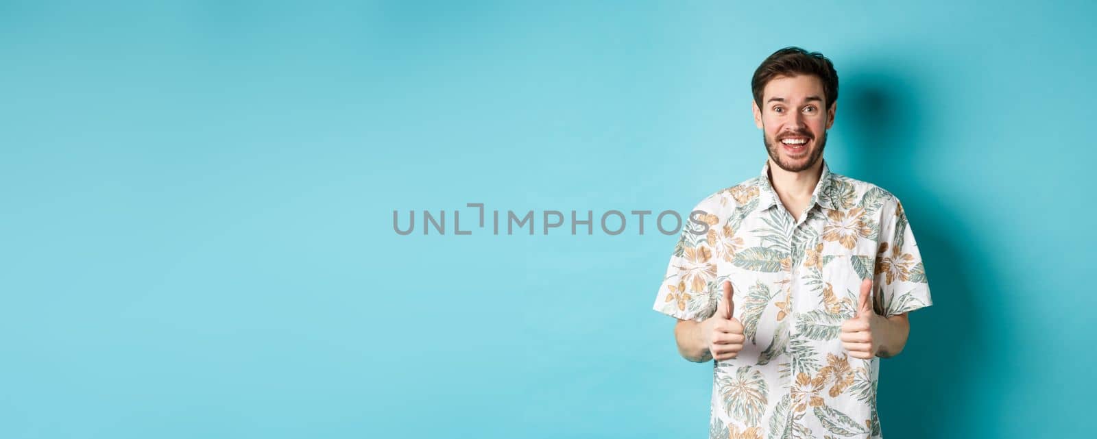 Summer holiday. Excited smiling tourist in hawaiian shirt show thumbs-up, praising hotel or vacation place, standing on blue background by Benzoix