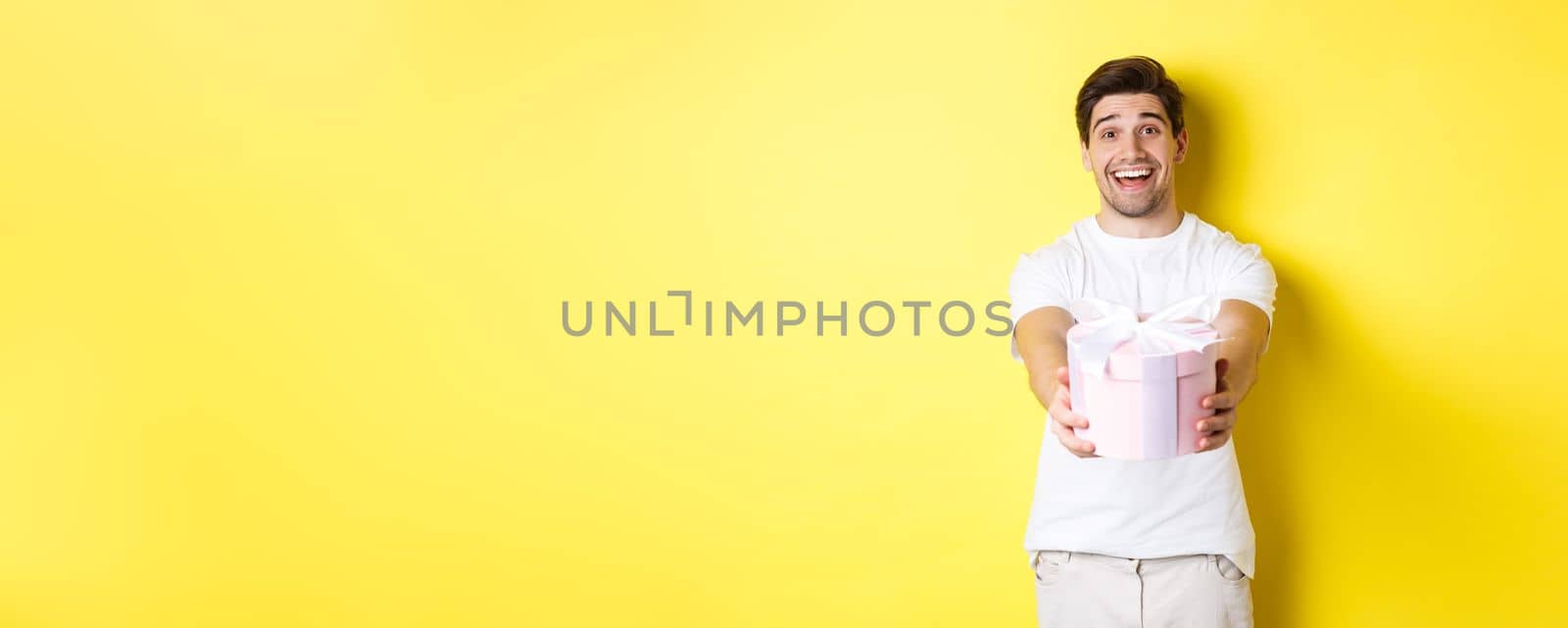 Concept of holidays and celebration. Smiling man giving you gift, congratulating, standing over yellow background with present.