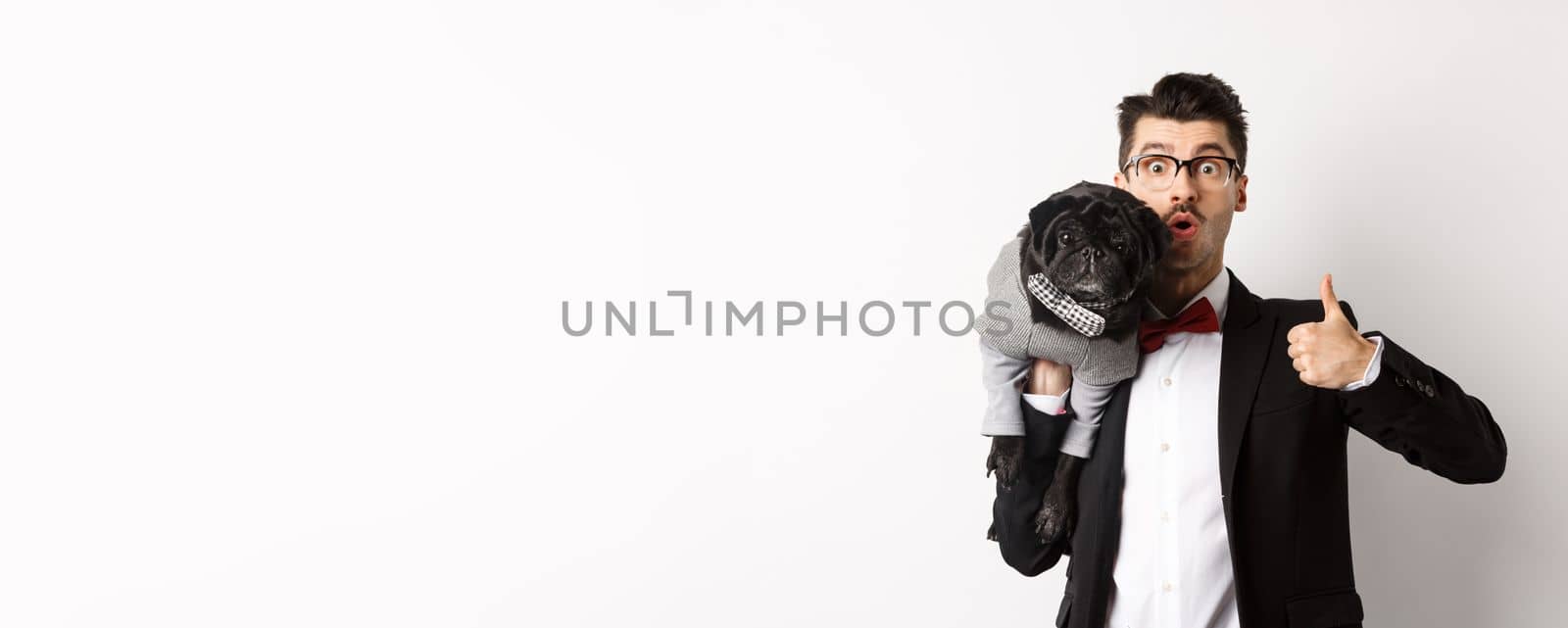 Happy young hipster in suit and glasses, showing thumb-up, holding cute black dog on shoulder, love his pug, standing over white background by Benzoix