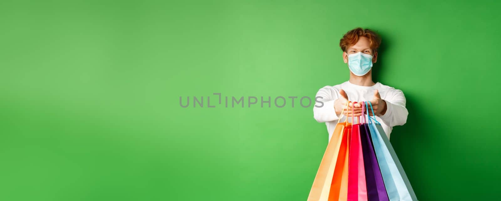 Happy young man in medical mask giving you shopping bags with purchases, smiling and wishing well, standing over green background. Covid-19 concept by Benzoix