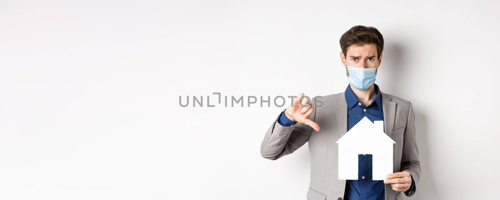 Real estate and covid-19 concept. Disappointed guy in medical mask and suit showing paper house cutout with thumbs-down, complaining on agency, white background by Benzoix