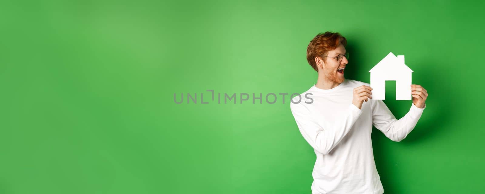 Real estate. Cheerful caucasian man with red hair, looking at paper house cutout and smiling amazed, buying property, standing over green background.