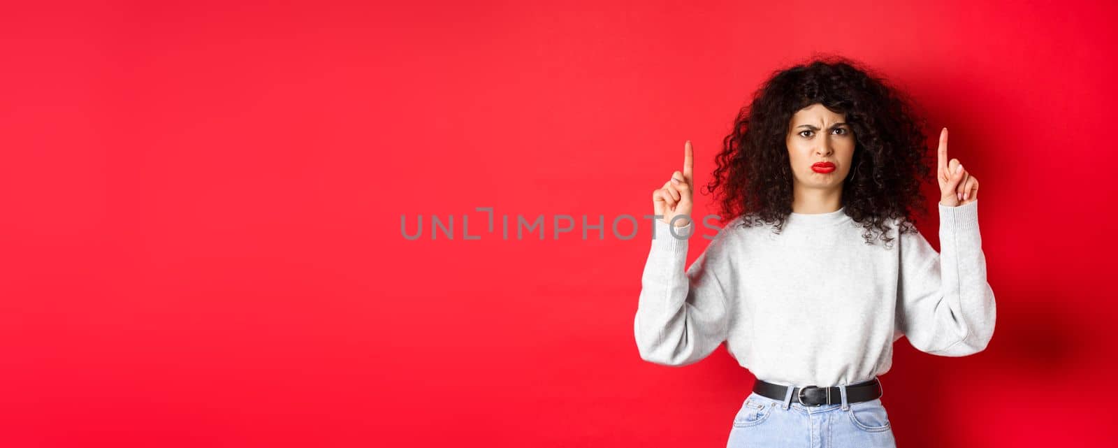 Grumpy young woman with curly hair frowning and grimacing unsatisfied, pointing fingers up at something bad, complaining on company, red background.