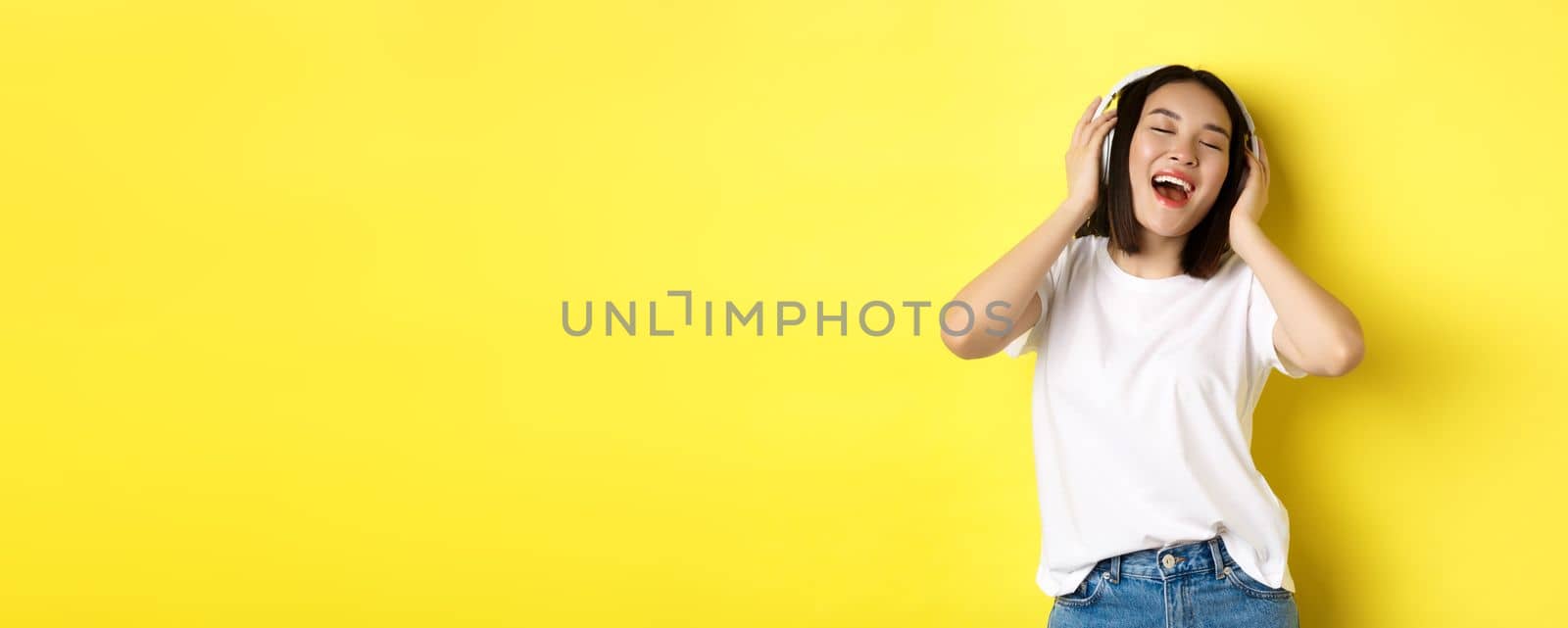 Attractive asian woman enjoying listening music in wireless headphones, smiling pleased and singing along, yellow background.