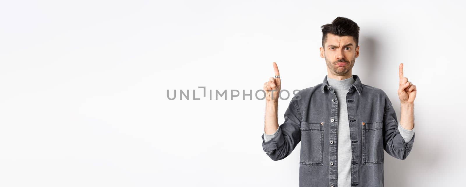 Confused funny guy with moustache pointing fingers up at something strange, frowning and pouting puzzled, standing on white background by Benzoix