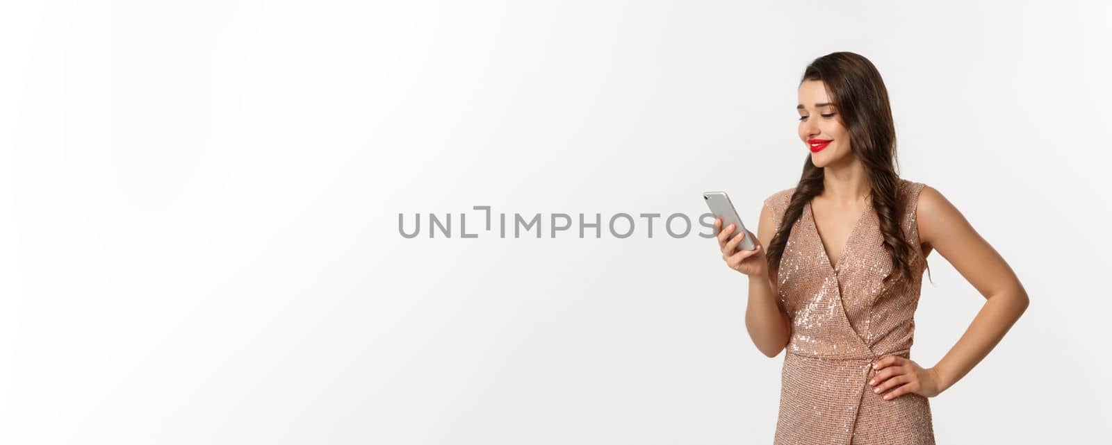 Christmas party and celebration concept. Happy young woman reading message on phone and smiling, using smartphone, wearing luxurious dress, white background.