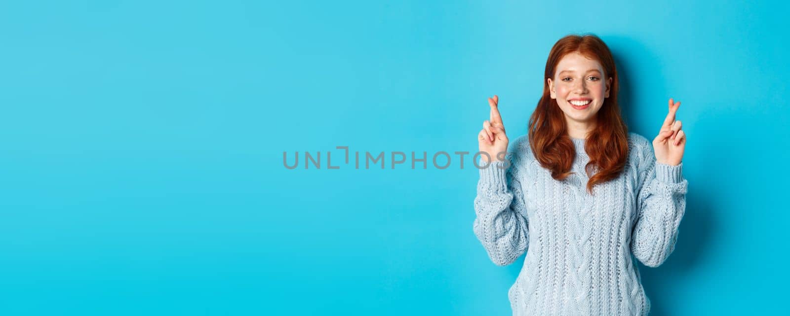 Hopeful redhead girl making a wish, cross fingers for good luck, smiling and anticipating good news or positive result, standing against blue background.