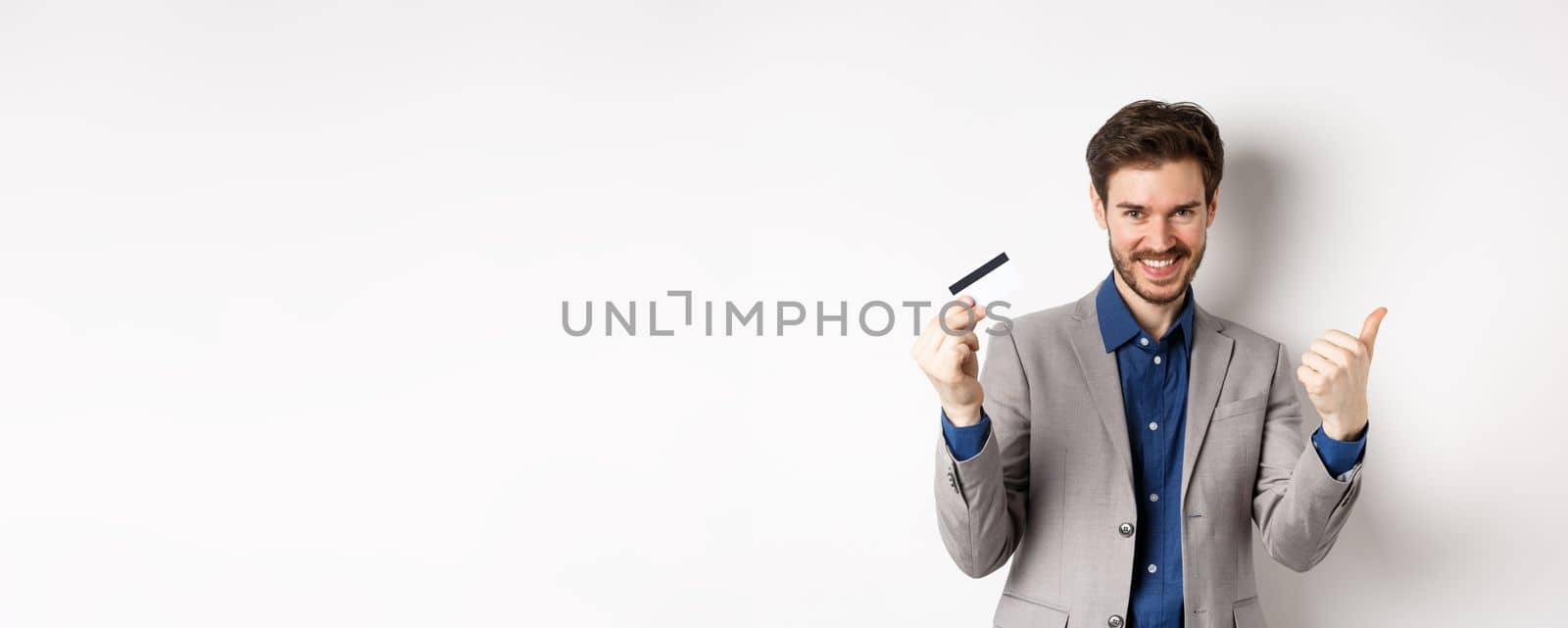 Happy bank client in business suit showing plastic credit card and thumb up, smiling satisfied, standing on white background by Benzoix