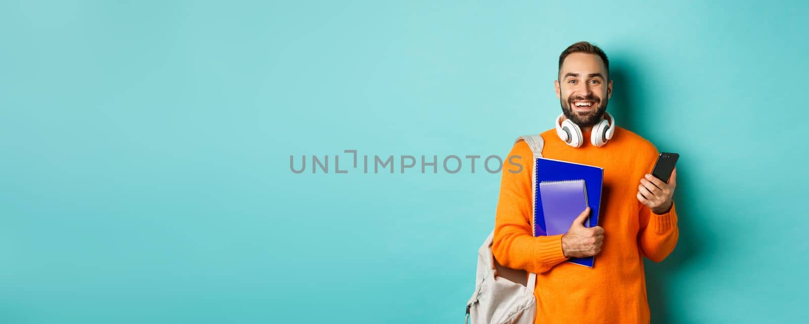 Education. Handsome male student with headphones and backpack, using smartphone and holding notebooks, smiling happy, standing over turquoise background.
