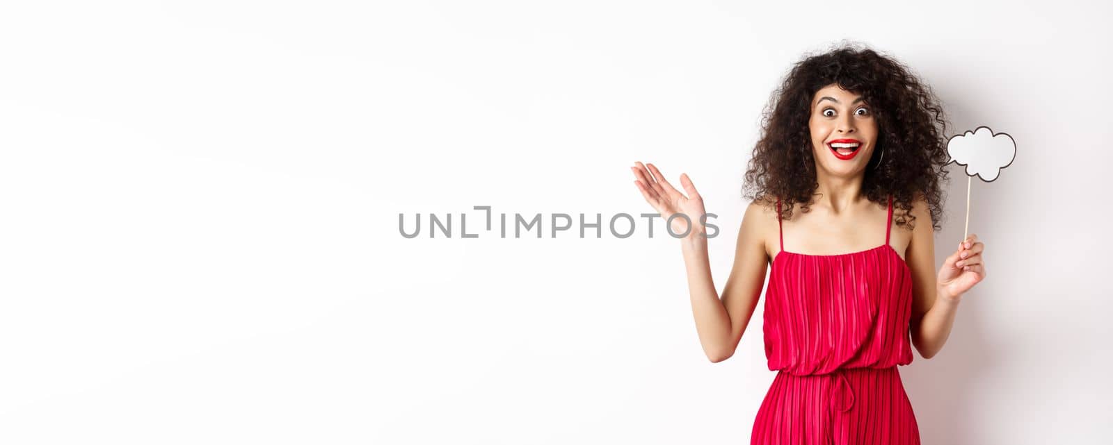 Happy young woman in red dress, holding comment cloud and looking surprised, standing over white background by Benzoix