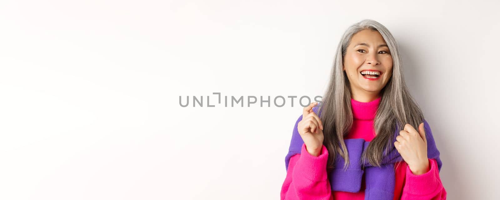Close-up of fashionable asian senior woman, laughing and having fun, looking left cheerful, standing over white background by Benzoix