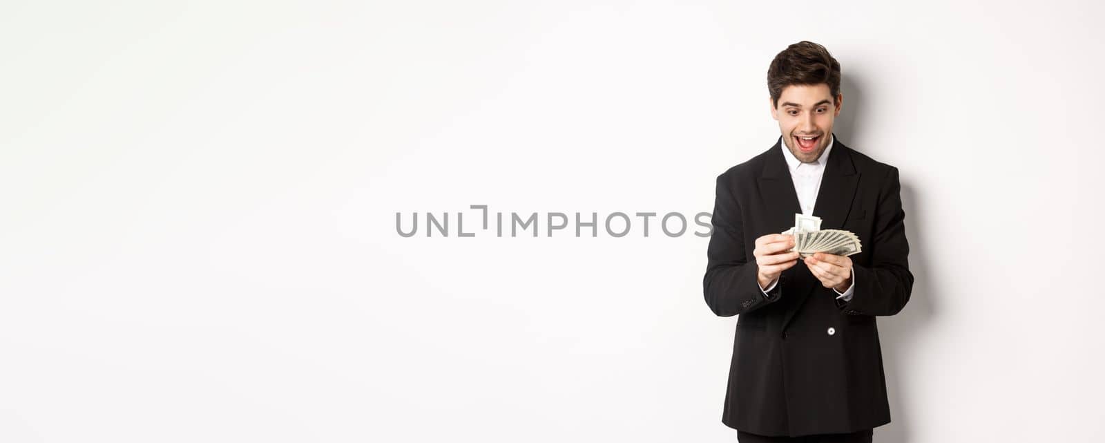 Image of excited handsome businessman, counting money and smiling amused, standing against white background in suit.
