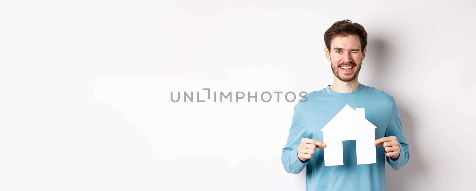 Real estate and insurance concept. Happy young man winking and smiling, showing paper house cutout, standing over white background.