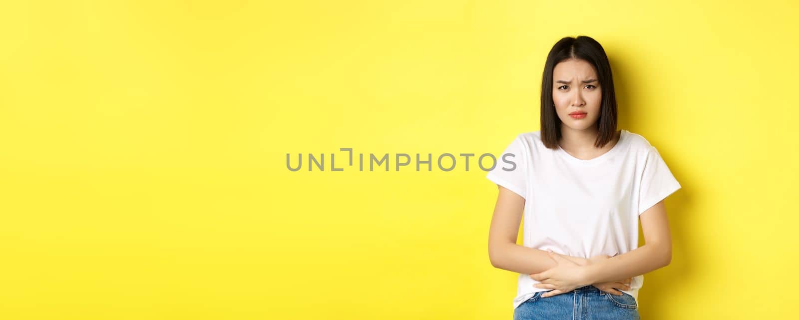 Image of asian woman feeling sick, having painful cramps, holding hands on belly and frowning from pain, discomfort during menstrual perion, standing over yellow background.