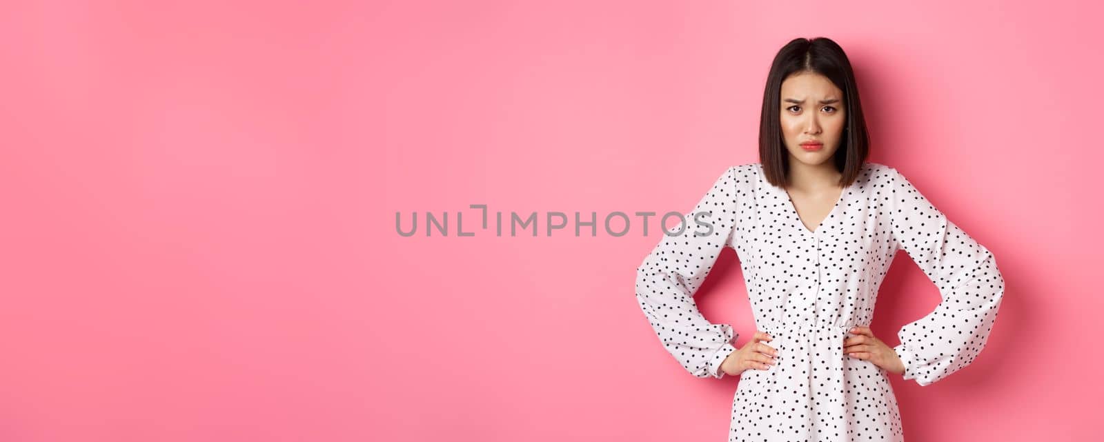 Cute offended girl frowning, holding hands on waist and staring at camera disappointed, standing upset and gloomy over pink background by Benzoix