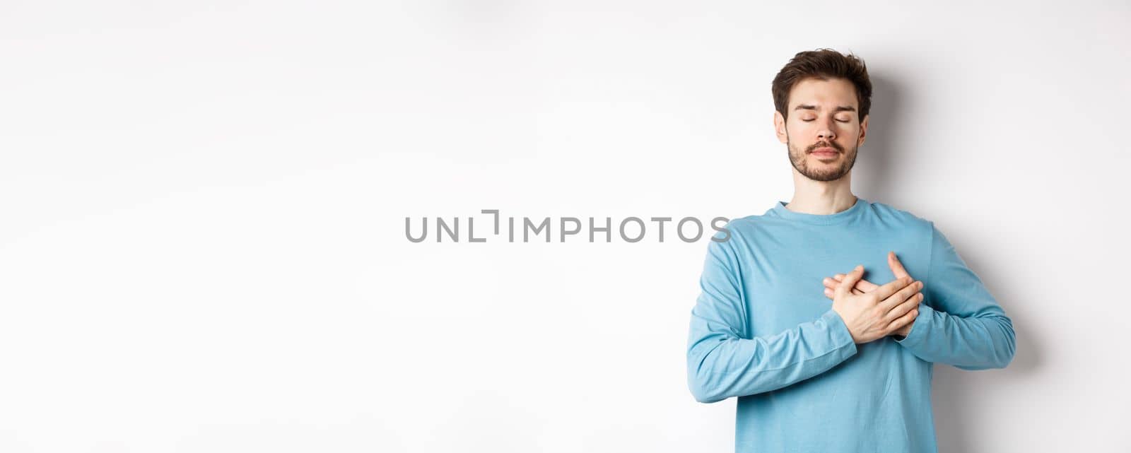 Portrait of young caucasian man with beard standing with hands on heart and closed eyes, dreaming about happy moments, standing over white background.