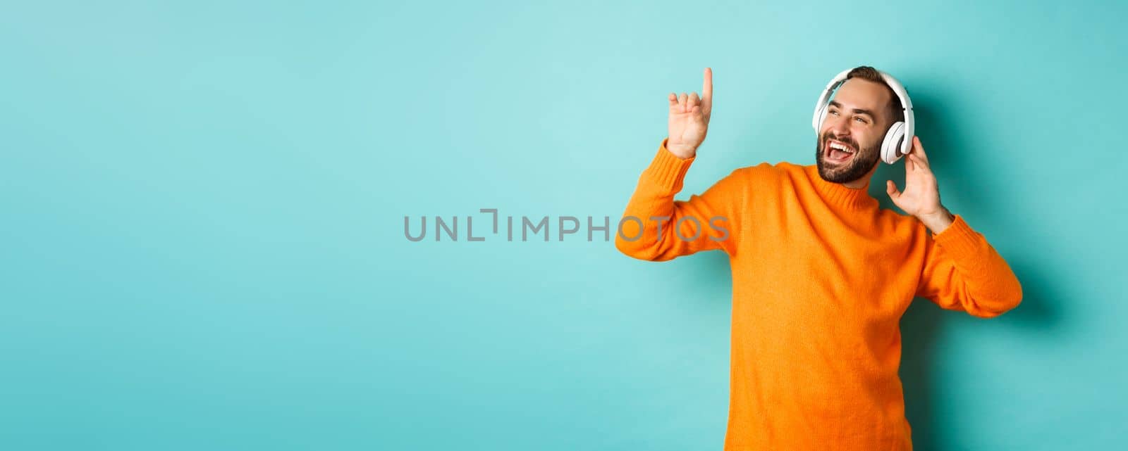 Happy adult man in orange sweater, looking up and listening music in headphones, standing over blue background.