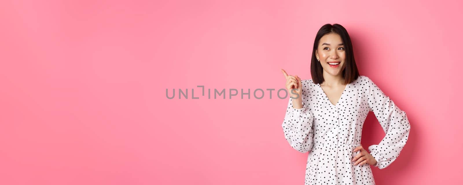 Happy asian girl in dress smiling satisfied, pointing finger at upper left corner, nod in approval, standing in dress against pink background by Benzoix