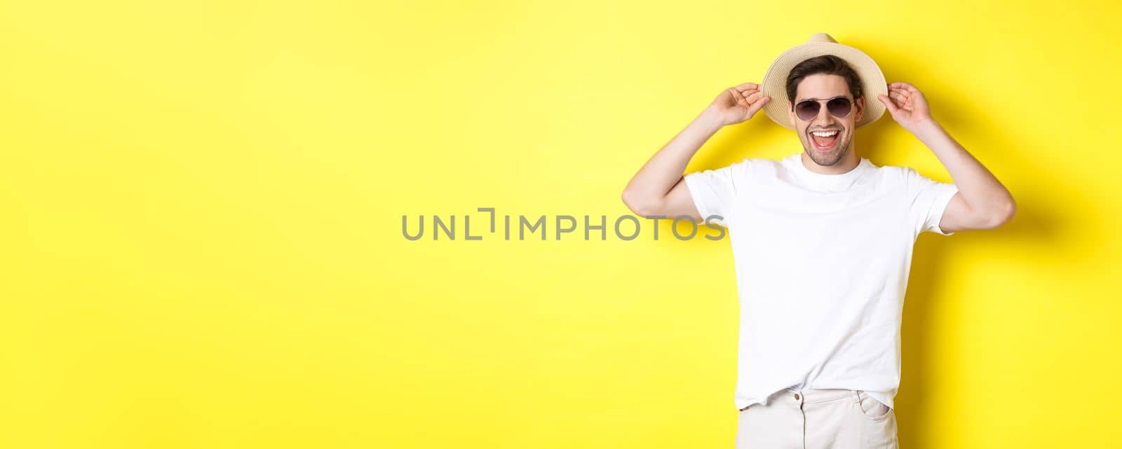 Happy man on vacation, wearing straw hat and sunglasses, smiling while standing against yellow background.