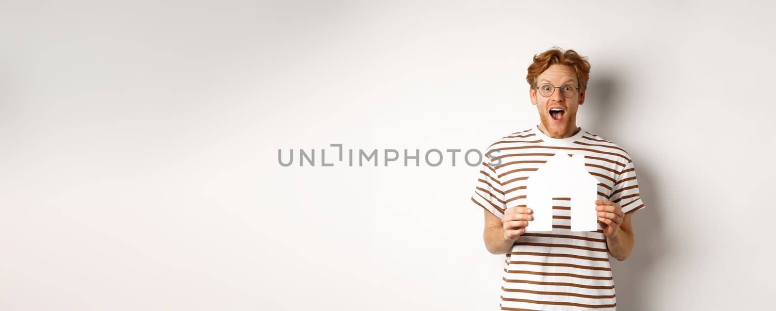 Happy and surprised redhead man winning house, holding paper home model and staring at camera, standing joyful over white background.