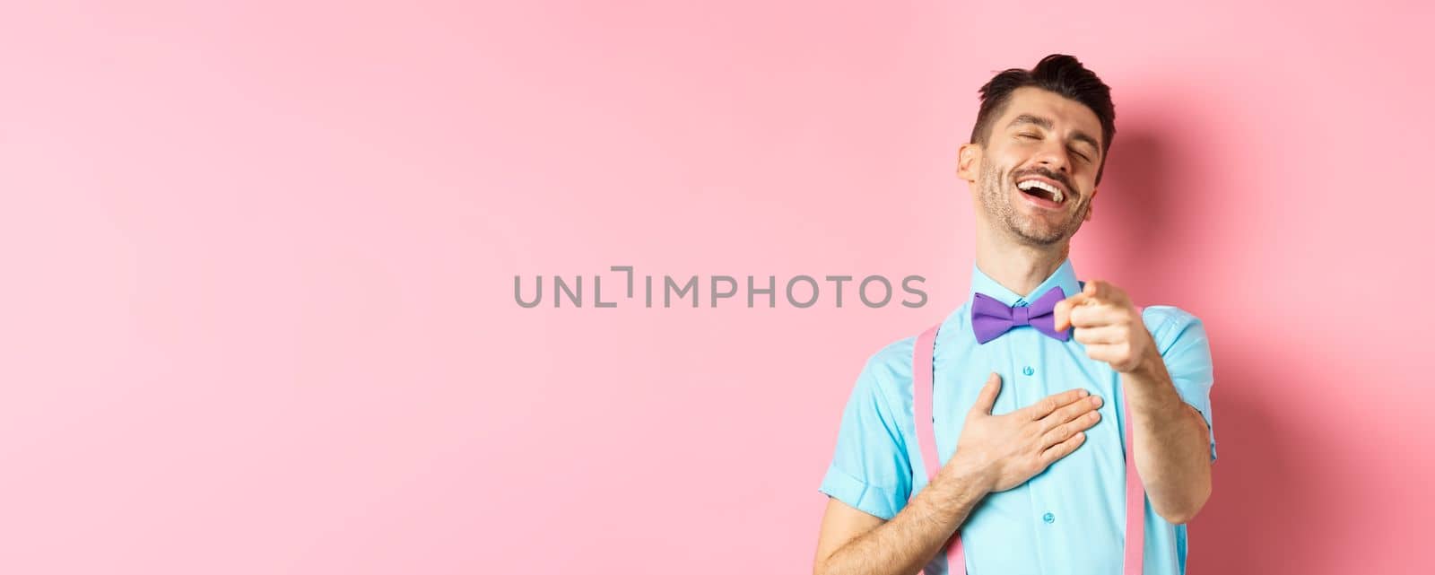 Handsome guy in bow-tie pointing finger at camera and laughing on something funny, checking out hilarious promo, standing on pink background.