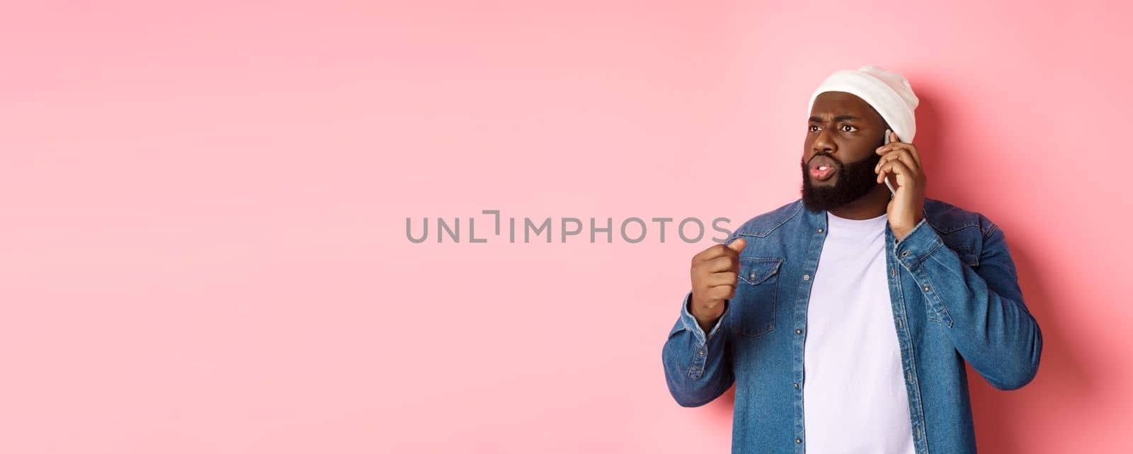 Angry african-american man talking on phone and threaten someone with raised fist, looking bothered at person, standing over pink background by Benzoix